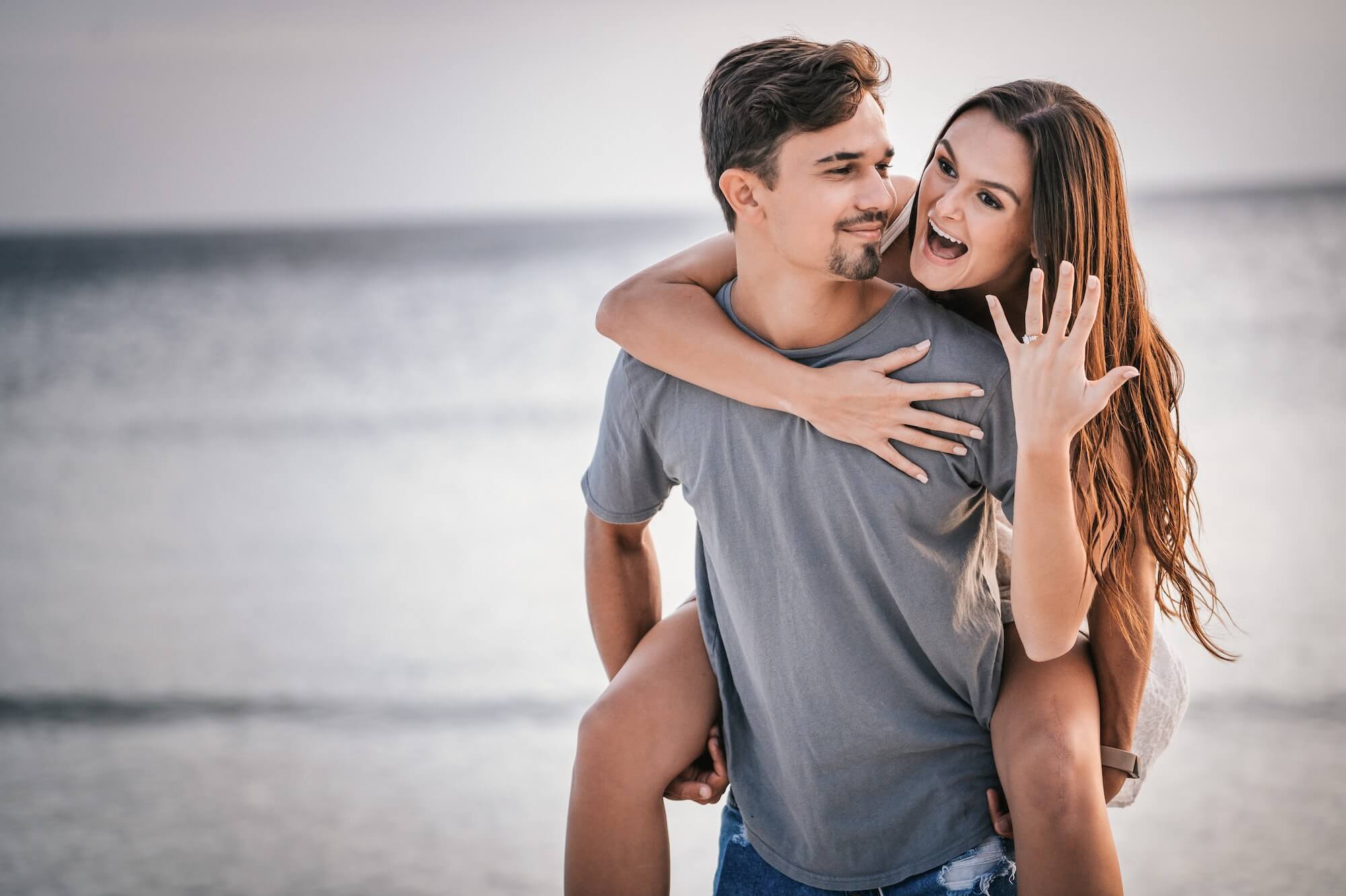 Happy couple on a beach, woman on man’s back showing off engagement ring.
